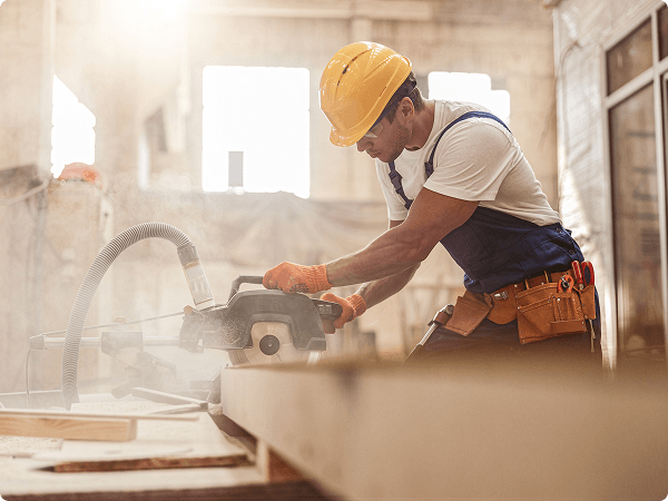 Construction worker wearing a yellow hard hat and tool belt operating a circular saw on a wooden beam in a workshop filled with natural light and wood dust, highlighting craftsmanship and manual labor.