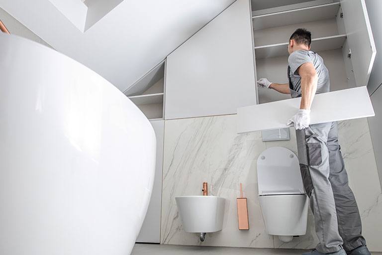 Contractor installing custom cabinetry in a modern bathroom featuring a freestanding bathtub, wall-mounted toilet, and sleek marble finishes, illustrating a high-end bathroom remodel in progress.