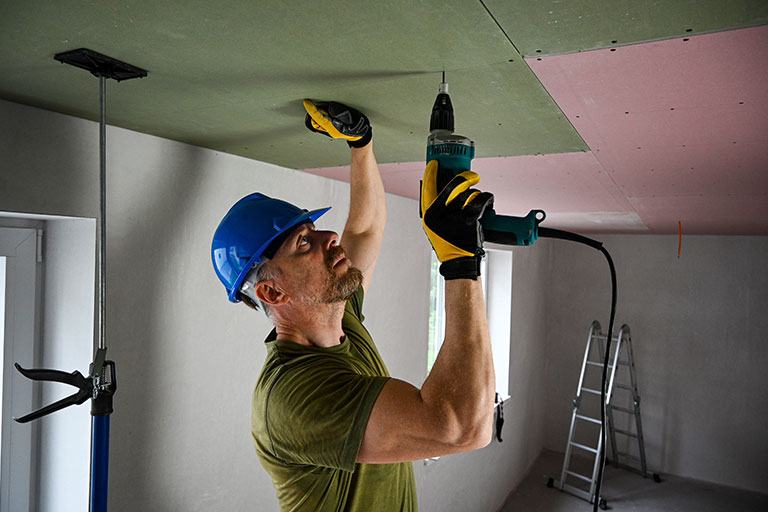 Construction worker in a blue hard hat using a power drill to install drywall panels on a ceiling, demonstrating skilled labor and precision work in an interior renovation.