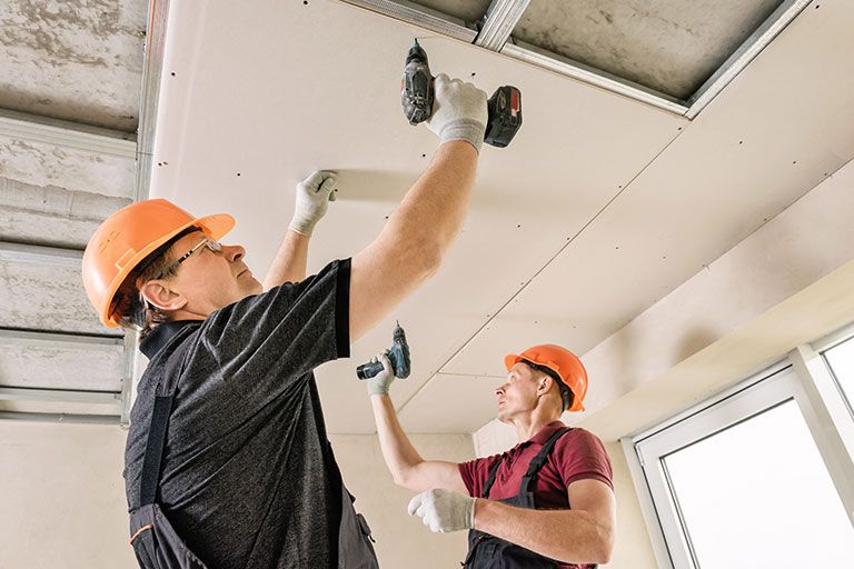 Two construction workers in orange hard hats and gloves installing drywall panels on a ceiling using power drills, highlighting teamwork and precision in drywall services.