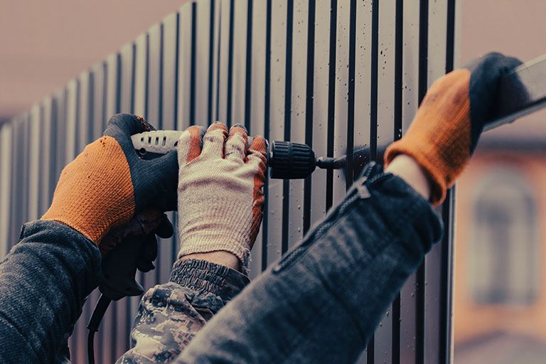 Close-up of gloved hands using a power drill to install vertical metal slats on a modern fence, highlighting precision craftsmanship and durable fencing installation.
