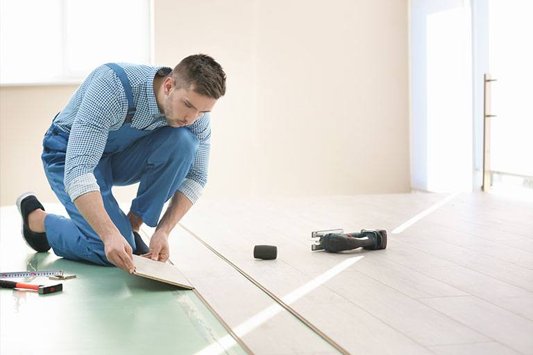 Construction worker in blue overalls kneeling while precisely placing a laminate plank during flooring installation in a bright, modern room, demonstrating craftsmanship and attention to detail.