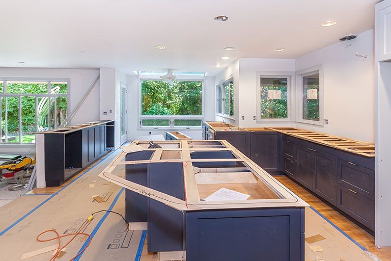 Modern kitchen undergoing renovation, with new dark cabinetry installed and wooden countertops being measured and fitted, surrounded by construction materials and tools in a bright space with large windows.