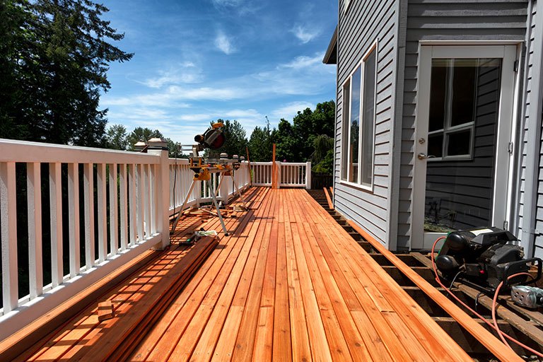 Spacious wooden outdoor deck under construction beside a gray-sided house, featuring white railing, power tools, and a clear blue sky—illustrating a professional deck installation project in progress.