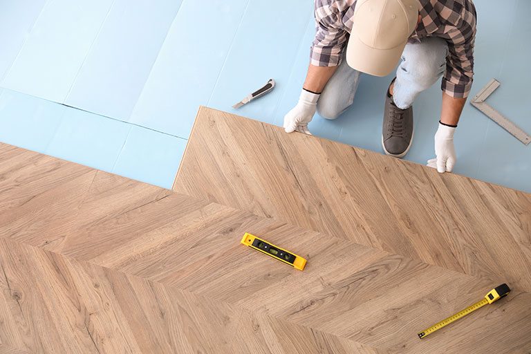 Overhead view of a contractor installing chevron-patterned wooden flooring with tools like a level and tape measure on a blue underlayment, emphasizing precision and design aesthetics.