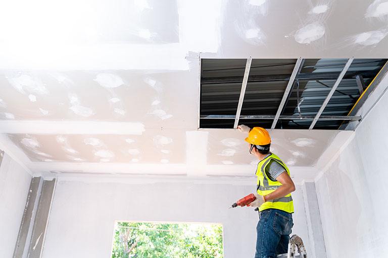 Worker wearing a yellow hard hat and safety vest inspecting a partially installed drywall ceiling with exposed metal framework, holding a drill while standing on a ladder in a bright room.