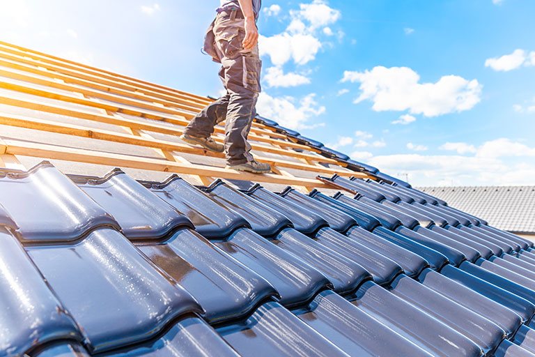 Roofer walking on a partially completed roof with glossy dark tiles under a sunny blue sky, highlighting professional roofing installation and repair in progress.