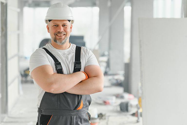 Smiling construction professional with folded arms and a white hard hat standing inside a renovation site, conveying trust, experience, and job site readiness.